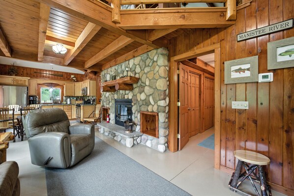 A wooden interior with a stone fireplace, leather sofa, and a dining area. - Camp Arrowhead: Northern Michigan Fall Retreat | Manistee Riverfront Cabin (Grayling)