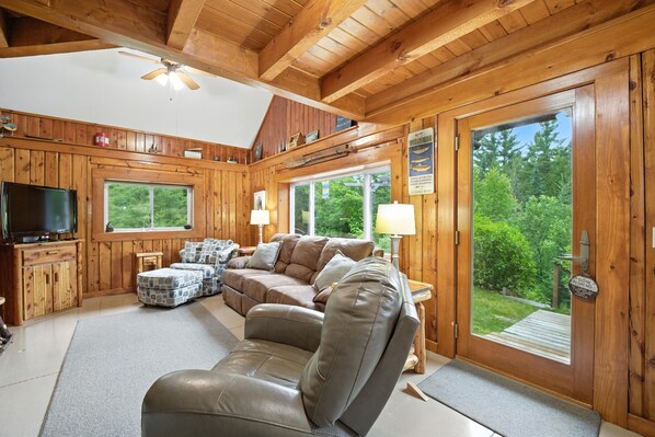 A wooden cabin interior with a sofa, television, and large windows overlooking greenery.