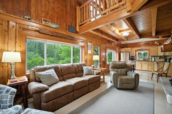 A cozy cabin interior with a wooden ceiling, a sofa, a recliner, and a kitchen area.