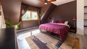 Une chambre à coucher avec un lit, un ventilateur de plafond, une fenêtre avec des rideaux, une table de chevet avec une lampe et une plante.