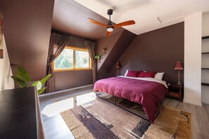 Un dormitorio con cama, ventilador de techo, ventana con cortinas, mesita de noche con lámpara y una planta.
