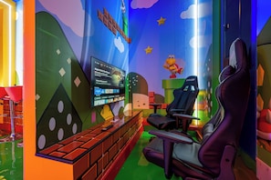 Une salle de jeux équipée d’un grand écran, de fauteuils de jeu et de décorations murales colorées.