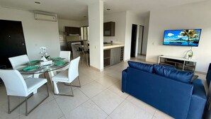 A modern living room with a blue sofa, a dining table with white chairs, and a kitchen in the background. - Deluxe 2 Bed 2 Bath Condo with a (Large outdoor Pool/Hot Tub) (Cancún)