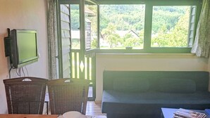 Une pièce avec un canapé vert, une table avec un set de table, une chaise en osier, un téléviseur fixé au mur et une fenêtre donnant sur un paysage verdoyant.