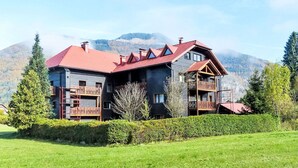 Un grand bâtiment à plusieurs étages, doté d’un toit rouge et de balcons en bois, entouré d’une pelouse bien entretenue et d’arbres, le tout se dressant sur un fond de montagnes.