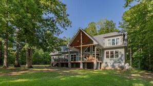 A large, two-story house with a prominent wooden beam structure on the second floor, surrounded by lush greenery. - Lakefront: Hot tub, Dock, Firepit, Kayaks, Grill, Sleeps 13 (Henrico)