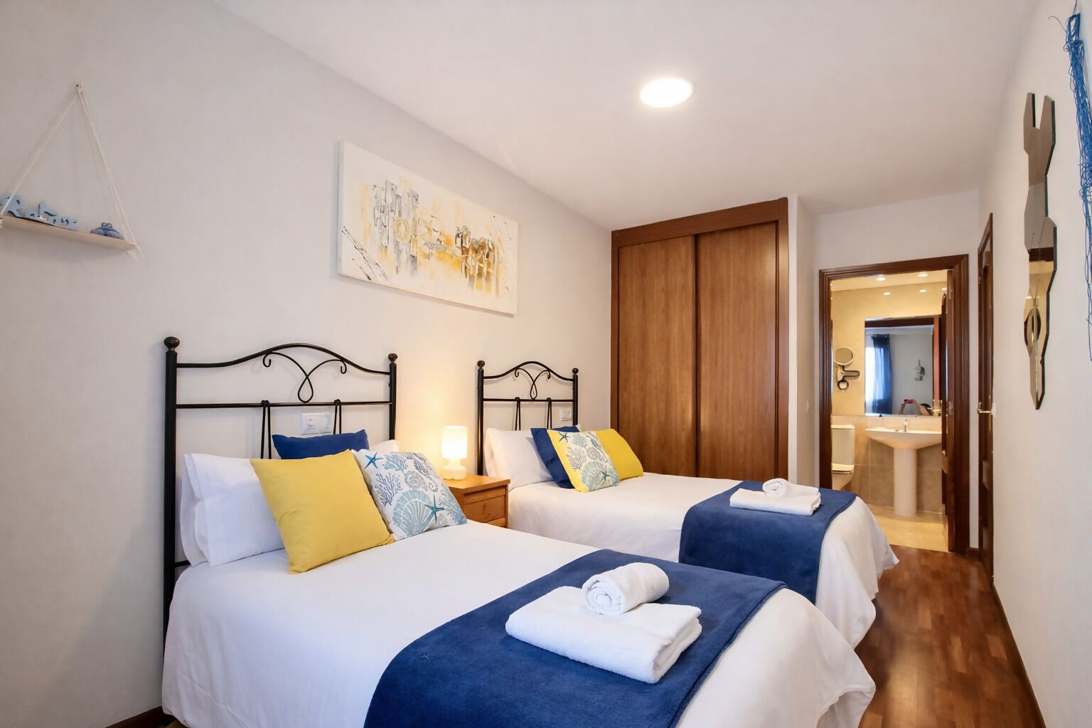 Habitación de hotel con dos camas, un armario de madera, un baño visible a través de una puerta abierta y una obra de arte colgada en la pared.