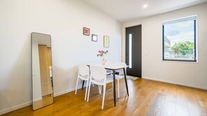 Una habitación con piso de madera, una mesa blanca con dos sillas, un espejo y una ventana con vistas a árboles y edificios.