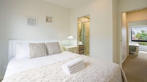 Habitación de hotel con cama, mesita de noche, lámpara, baño con ducha de vidrio y balcón con vistas.