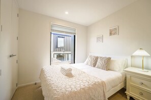 A bedroom with a bed, bedside table, lamp, and a window with a view of a balcony.