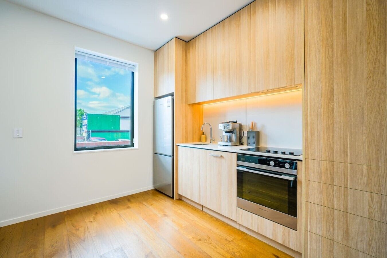 A modern kitchen with wooden cabinets, a built-in oven, and a window with a view of a green building.