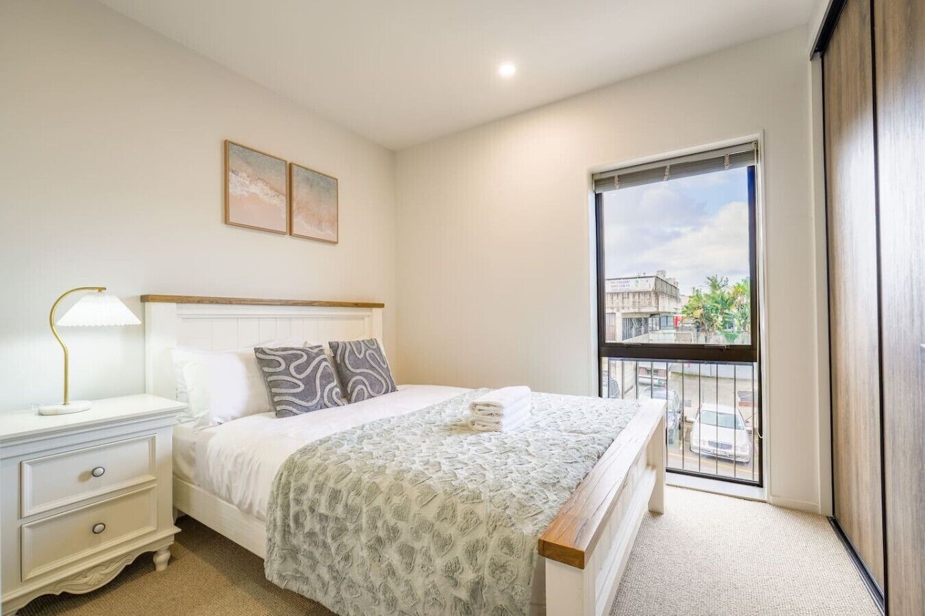 A bedroom with a bed, bedside table, lamp, and a view of the outside through a window.