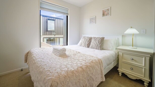 A bedroom with a large bed, a bedside table with a lamp, and a window with blinds. - Chic 2-Bedroom Retreat in Grey Lynn Minutes to CBD (Auckland)