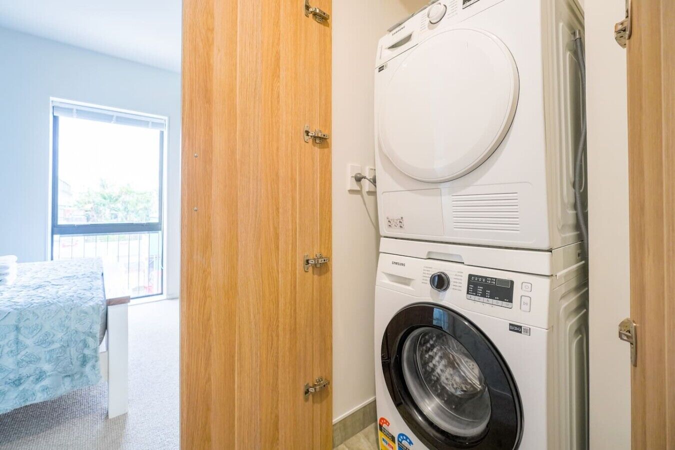 A laundry area with a front-loading washer and dryer, a wooden door, and a bed with a blue cover.