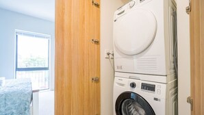 A laundry area with a front-loading washer and dryer, a wooden door, and a bed with a blue cover. - Chic 2-Bedroom Retreat in Grey Lynn Minutes to CBD (Auckland)