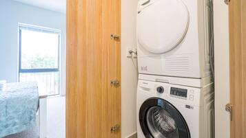 A laundry area with a front-loading washer and dryer, a wooden door, and a bed with a blue cover.
