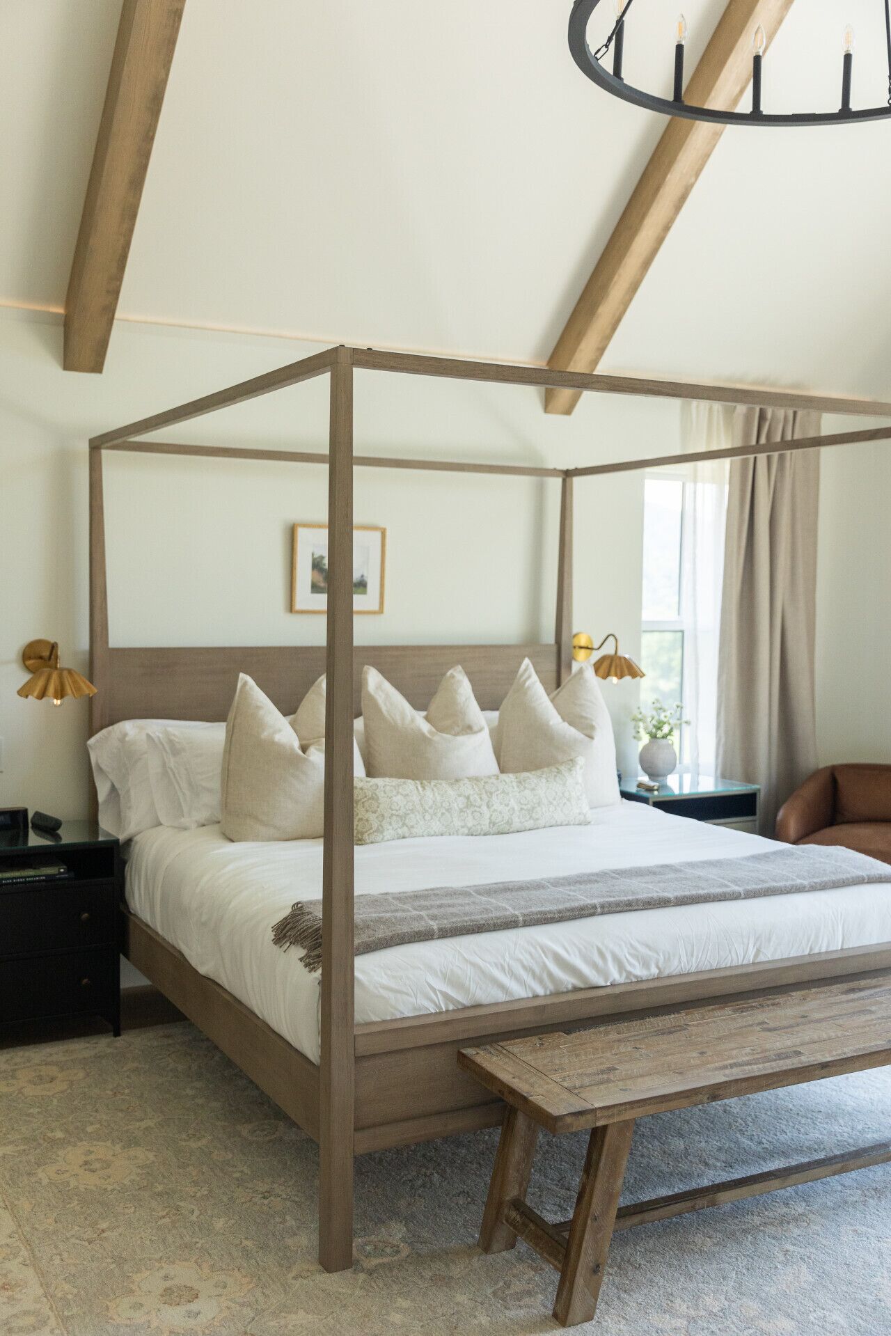 A four-poster bed with a wooden frame and white bedding, a bedside table with a lamp, and a view of the outdoors through a window.