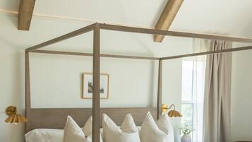 A four-poster bed with a wooden frame and white bedding, a bedside table with a lamp, and a view of the outdoors through a window.