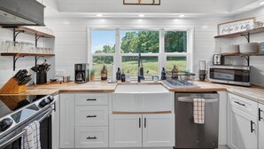 A modern kitchen with white cabinetry, a large window offering a view of a green landscape, and various kitchen appliances and utensils.