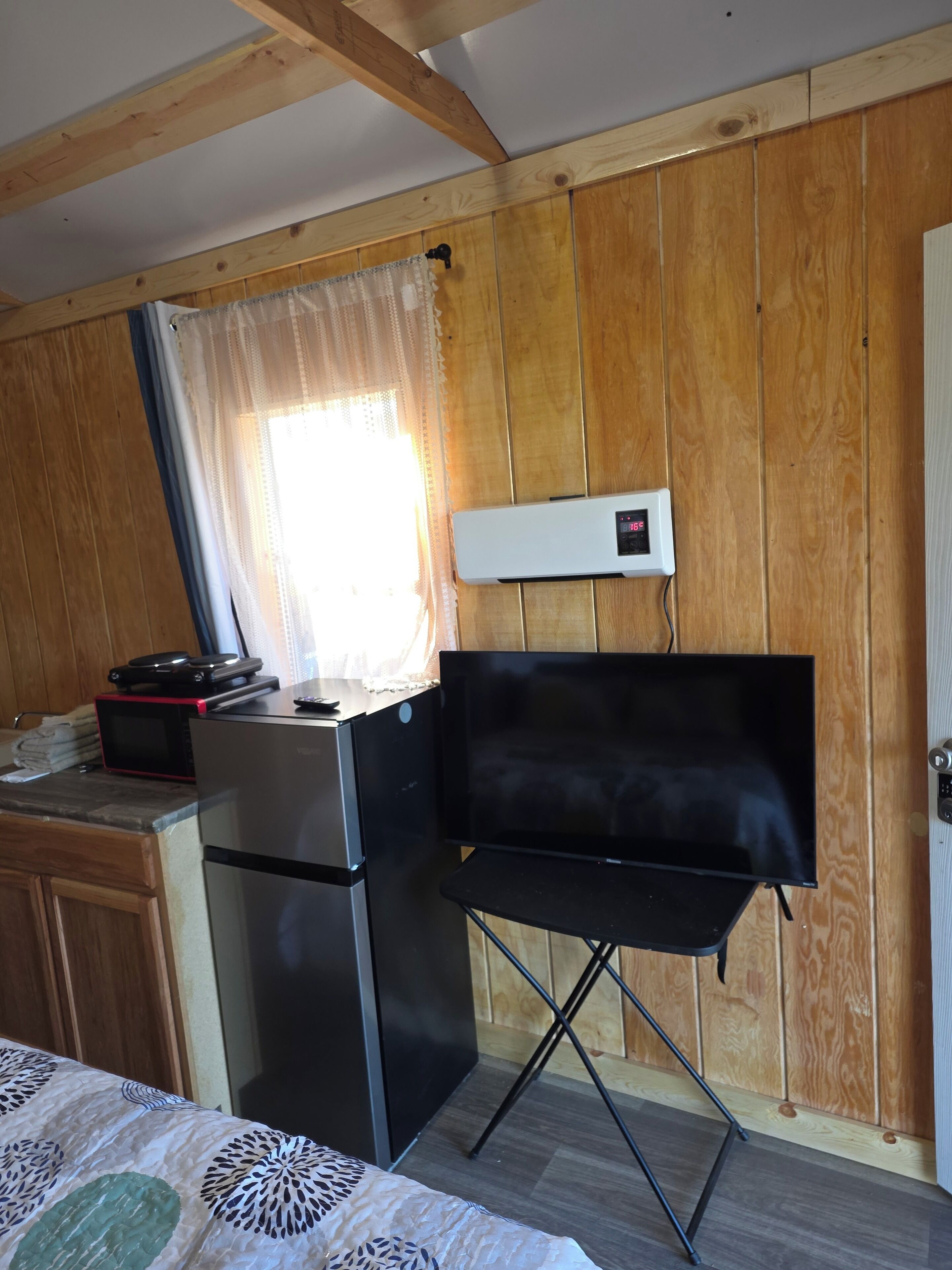 Una kitchenette compacta con refrigerador, microondas y una pequeña encimera. Hay una silla plegable y un televisor de pantalla plana montado en la pared.