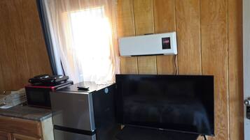 Una kitchenette compacta con refrigerador, microondas y una pequeña encimera. Hay una silla plegable y un televisor de pantalla plana montado en la pared.