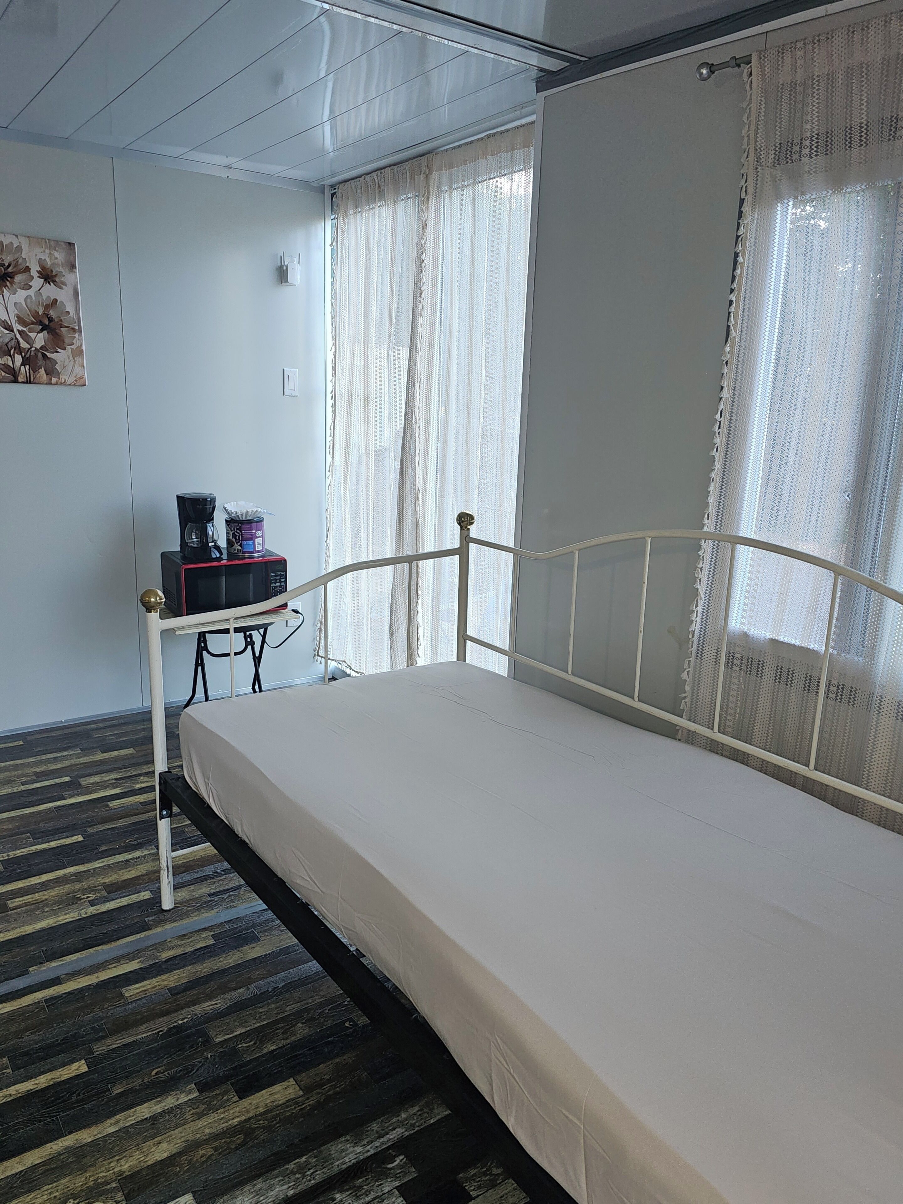 A single bed with a metal frame, a small table with a coffee maker, and a painting on the wall.