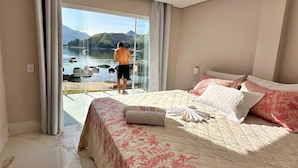 A hotel room with a bed, a large window with a view of a lake and mountains, and a balcony with a person standing outside. - Mansion on the sand, private beach, heated jacuzzi, wet bar (Ponta do Sapê Angra dos Reis)