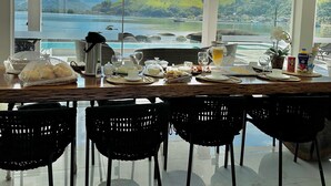 A dining area with a view of a lake and mountains, featuring a table set with food and drinks, black chairs, and pendant lights. - Mansion on the sand, private beach, heated jacuzzi, wet bar (Ponta do Sapê Angra dos Reis)