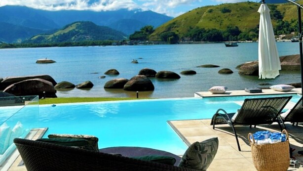 A poolside area with a view of a lake and mountains. There are lounge chairs and a white umbrella. - Mansion on the sand, private beach, heated jacuzzi, wet bar (Ponta do Sapê Angra dos Reis)