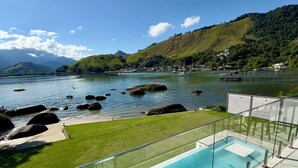 A swimming pool with a clear view of the surrounding landscape, including a body of water, rocks, and distant hills. - Mansion on the sand, private beach, heated jacuzzi, wet bar (Ponta do Sapê Angra dos Reis)