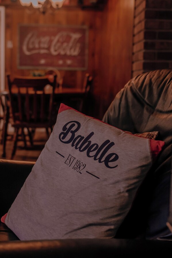 A pillow with the text 'Babelle EST 1982' on a couch in a room with a brick wall and a Coca-Cola sign in the background.