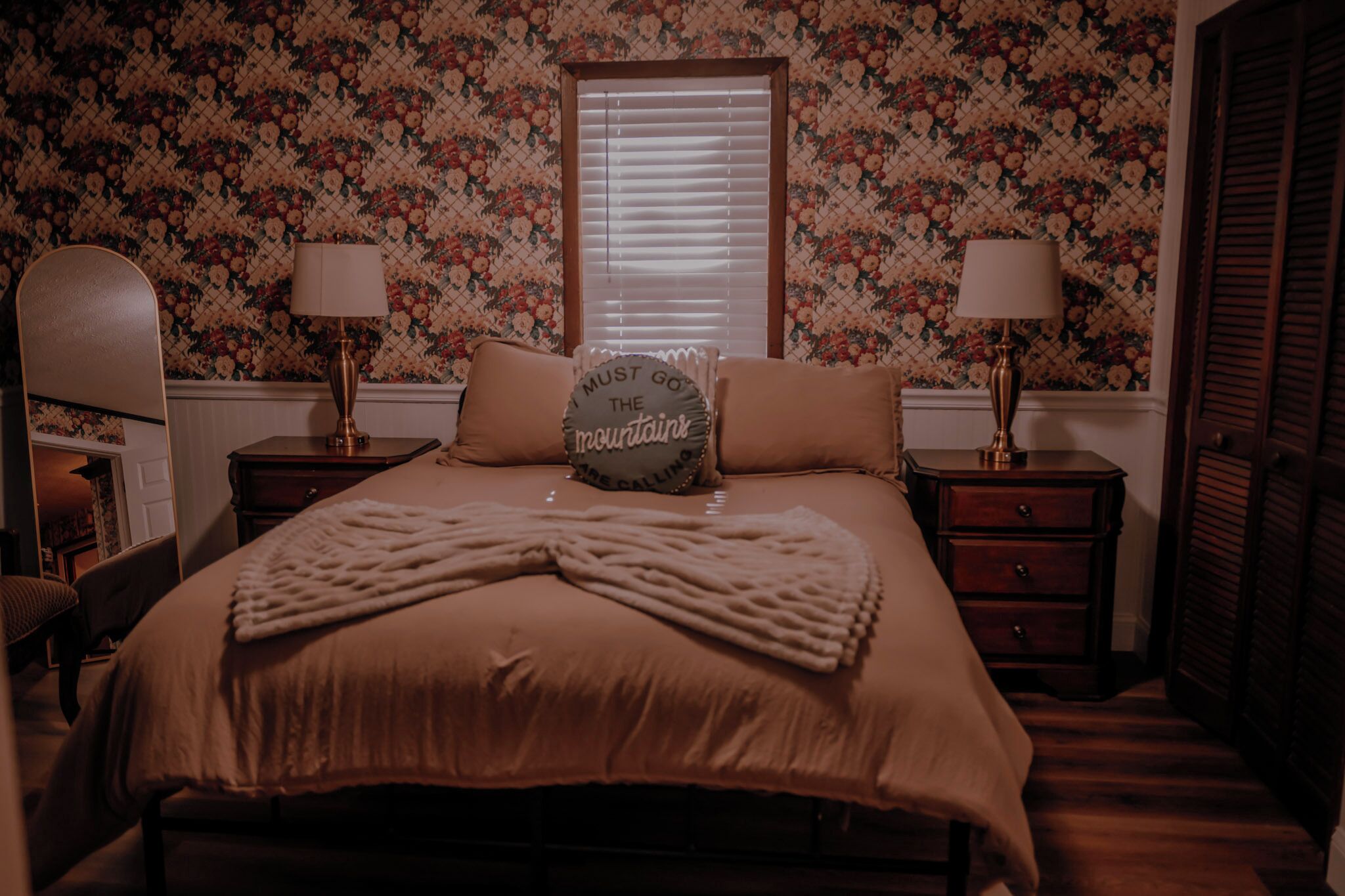 A bedroom with a floral wallpaper, a bed with a mountain-themed pillow, two bedside lamps, and a wooden floor.