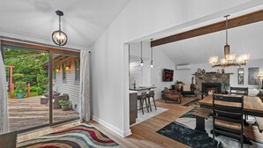 A spacious living area with a wooden dining table, a fireplace, and a large window overlooking a deck. - Near pool & beach. Private Hot Tub. AC, Firepit (Pocono Lake)