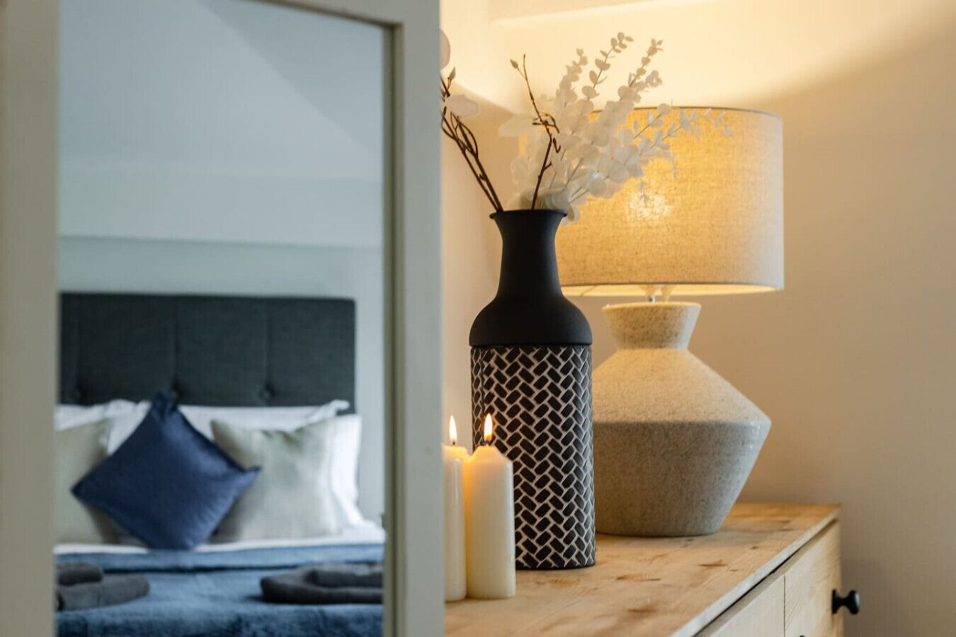 A neatly made bed with a dark headboard, a bedside table with a lamp, a vase with branches, and a lit candle.