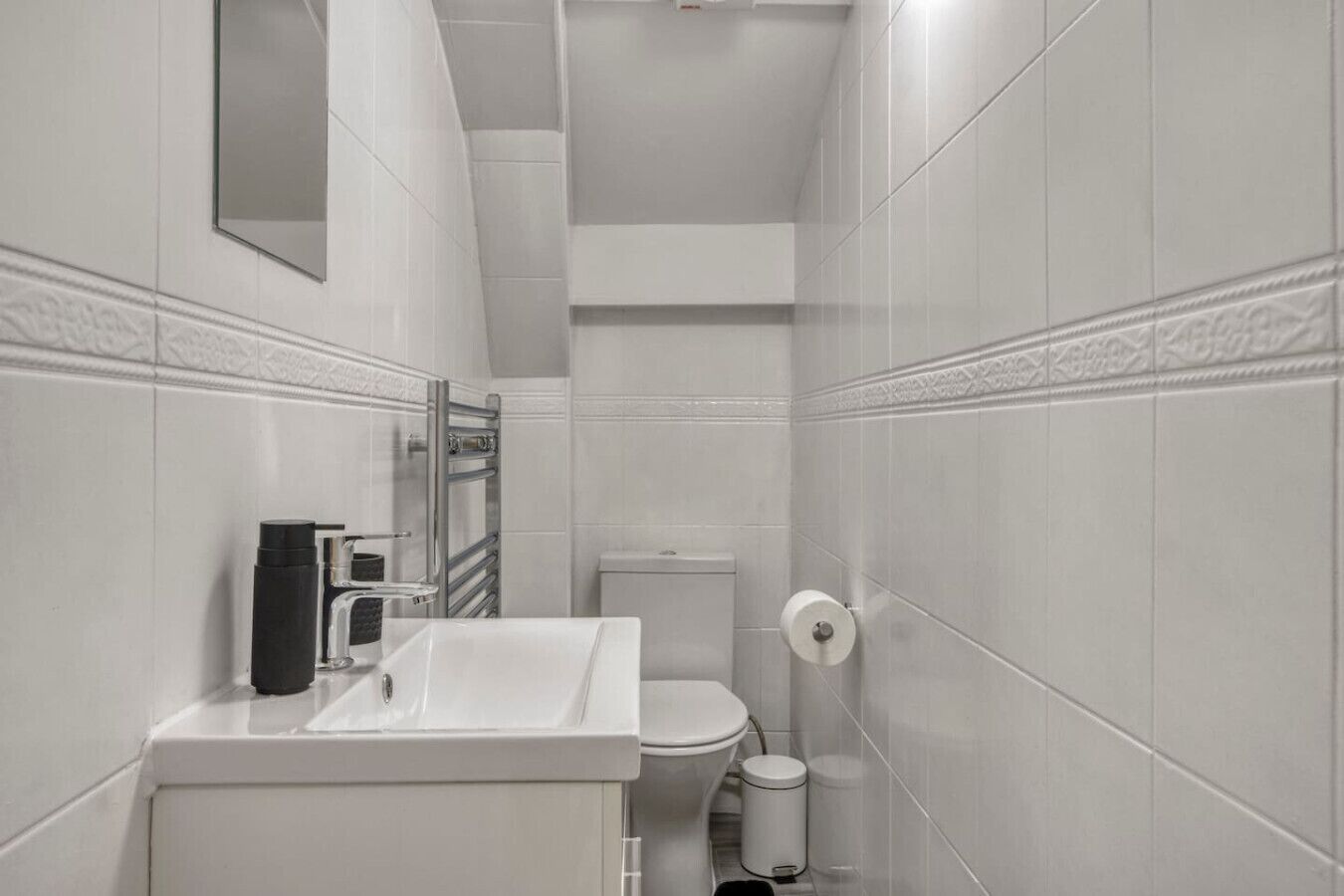 Un baño pequeño y moderno con azulejos blancos, un lavamanos blanco, un inodoro y un bote de basura.