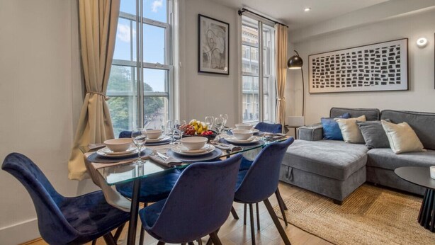 Un coin repas avec une table en verre dressée pour six personnes, des chaises bleues et un canapé gris en arrière-plan.