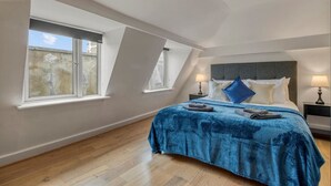 Une chambre avec un plafond en pente, un grand lit recouvert d’une couverture bleue, deux tables de chevet avec des lampes et deux fenêtres.
