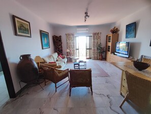 Un salon comprenant un canapé, deux fauteuils en osier, un téléviseur, un grand vase et un tableau accroché au mur.
