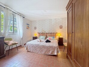 A bedroom with a large bed, two bedside tables with lamps, a wardrobe, a chair, and a window with curtains.