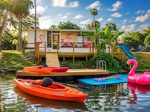 A dock with kayaks, a flamingo float, and a building with a deck and pool. - Fun-Filled Flamingo Getaway | Kayaks, Water Slide, Patio & Fully Fenced Waterfront Yard (Weeki Wachee)