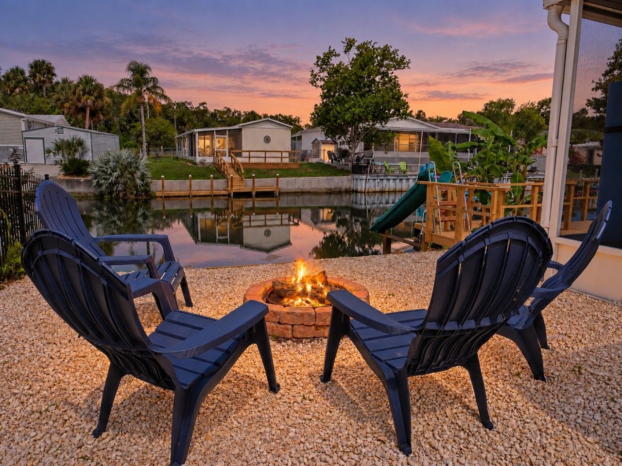 A fire pit with two chairs facing it, surrounded by gravel, with a view of a water body and houses in the background.