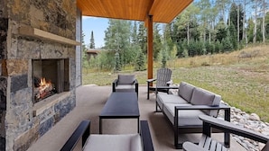 Un patio al aire libre con chimenea de piedra, zona de estar y vistas a un promontorio boscoso.