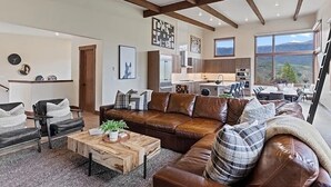 Amplia sala de estar con un sofá grande de cuero marrón, una mesa de centro de madera y una chimenea. Se observa una cocina al fondo con electrodomésticos modernos y un amplio ventanal con vistas al exterior.