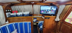 A compact interior with a television displaying various channels, a small table with items, and a bed with striped bedding. - Nautical Nights - House Boat (Seward)