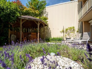 Une terrasse couverte, aménagée avec du mobilier d’extérieur, entourée d’un jardin où poussent des plants de lavande et bordée d’un chemin en pierre.