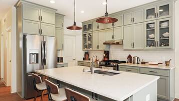 A kitchen with light green cabinets, a stainless steel refrigerator, a large island with bar stools, pendant lighting, and a tiled backsplash.