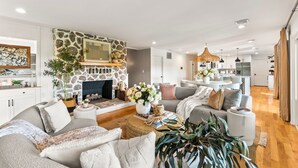 A modern living room with a stone fireplace, a sofa, a coffee table, and a dining area with a chandelier. - *NEW* Waterfront Treehouse - Sweeping Views on 2.5 Acres (Knoxville)