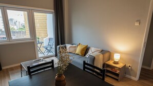 A modern living room with a dining table, a sofa with cushions, a small side table with a lamp, and a view of the outside through a window. - 3BR Flat by Casa da Música | Central, Sleeps 6 | D (Porto)