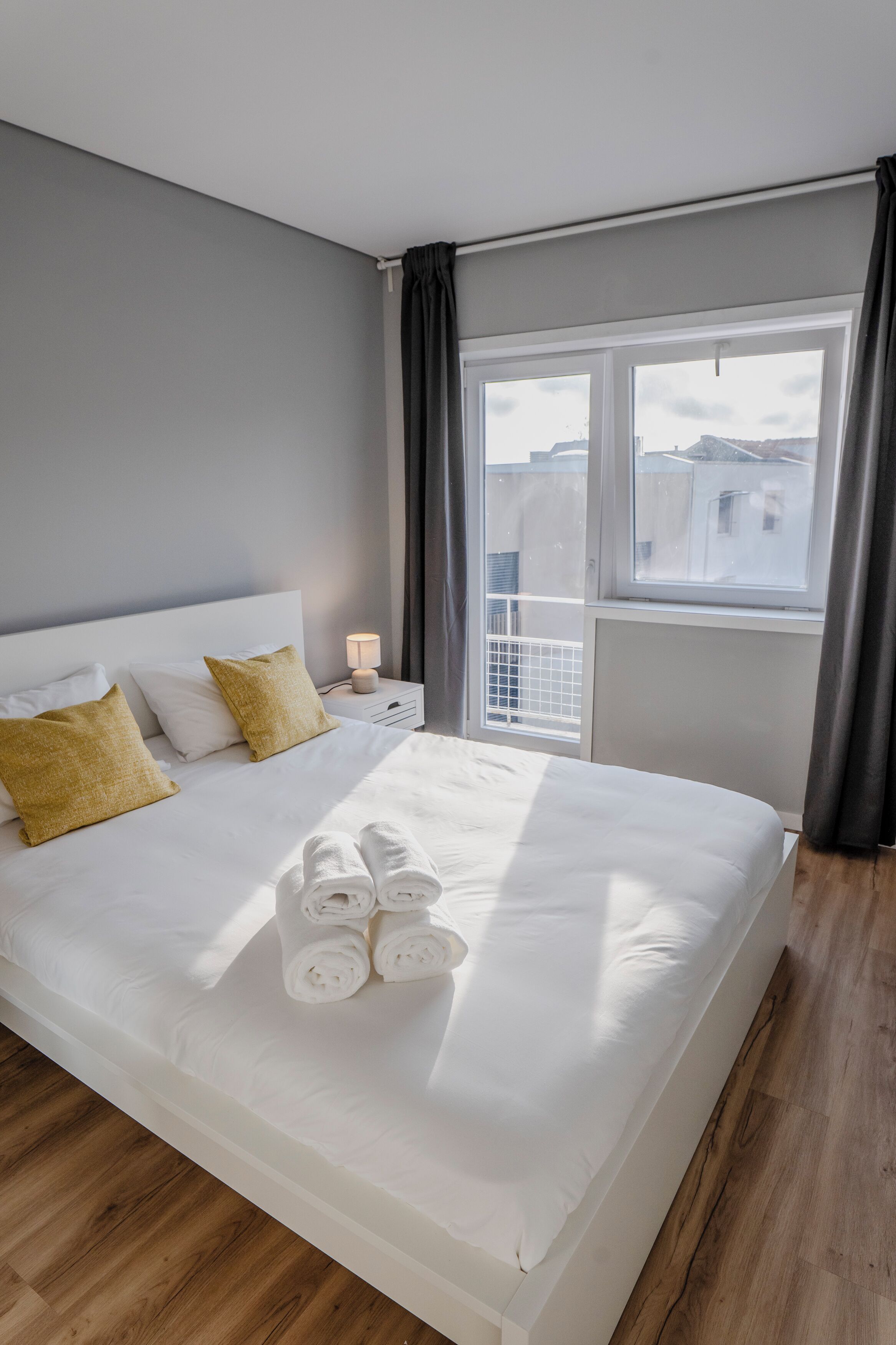 En pænt redt seng med hvide sengetæpper og sammenrullet håndklæder, et natbord med lampe, og et vindue med gardiner.