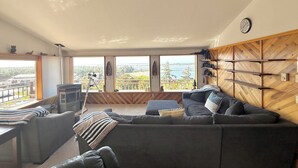 A spacious living room with a large gray sectional sofa, a wooden herringbone wall, and a view of the ocean through large windows.